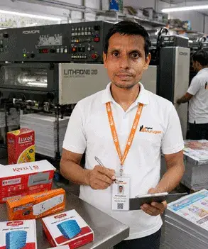 Pramod Mandal, Supervisor – Production at Digital Murugan, standing inside the printing and packaging production facility with a Komori offset printing machine, branded product boxes, official company polo shirt, and ID card.